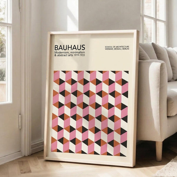 May include: Framed Bauhaus poster with geometric cube pattern in pink, orange, black and cream colors, displayed on light wooden floor next to beige upholstered chair. Natural light streams through white-framed windows in minimalist room setting. The artwork features three-dimensional cubic tessellation design with modernist typography at top.