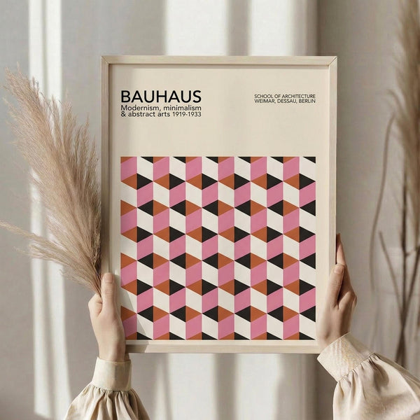 May include: Hands holding a framed geometric poster featuring a repeating cube pattern in pink, coral, black, and cream colors. The frame is light wood against a neutral background with dried pampas grass visible to the left. The poster displays "BAUHAUS" text at the top with dates 1919-1933 and references to modernism, minimalism and abstract arts.