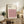 May include: Framed geometric poster with pink, white, and black cube pattern displayed on wooden sideboard next to fern plant, ceramic vase, and books in neutral living room setting with beige chair visible