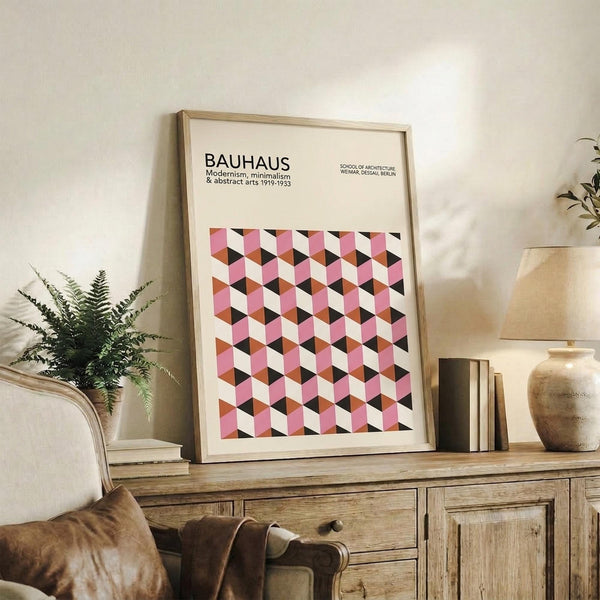 May include: Framed geometric poster with pink, white, and black cube pattern displayed on wooden sideboard next to fern plant, ceramic vase, and books in neutral living room setting with beige chair visible