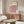 May include: Modern living room with wooden sideboard displaying a framed geometric poster featuring pink, coral, and black cube pattern. White upholstered chair, glass oval coffee table, and floor lamp visible. Poster shows three-dimensional hexagonal design in warm tones against cream background with black text header.