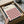 May include: Hands unboxing a framed geometric poster from cardboard packaging. The artwork features a repeating diamond pattern in pink, orange, black and cream colors with "BAUHAUS" text at top. Black frame sits among white tissue paper with tape roll visible nearby.