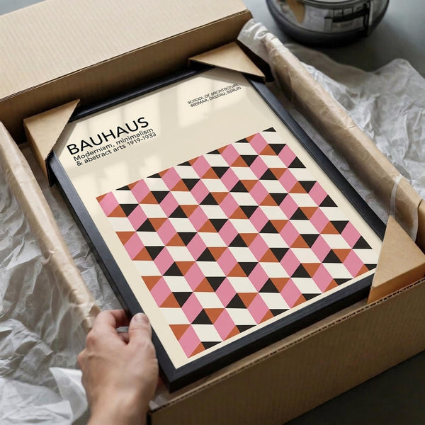 May include: Hands unboxing a framed geometric poster from cardboard packaging. The artwork features a repeating diamond pattern in pink, orange, black and cream colors with "BAUHAUS" text at top. Black frame sits among white tissue paper with tape roll visible nearby.