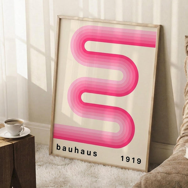 May include: Framed poster with pink gradient wavy ribbon design against cream background, displaying "bauhaus 1919" text below. Light wood frame leaning against white wall corner next to beige textured rug, wooden side table with white coffee cup, and tan fabric in soft natural lighting.