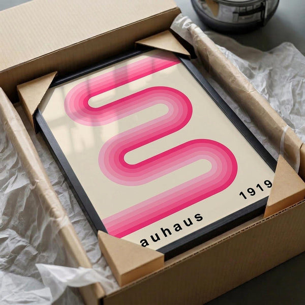 May include: Pink ribbon-shaped design with gradient shading on cream background, displayed in black frame. Artwork shows "auhaus 1919" text in black letters. Frame sits in opened cardboard shipping box with white tissue paper padding. Wooden dowels for rolling artwork visible alongside. Scene photographed on white surface with partial view of dark bowl in background.