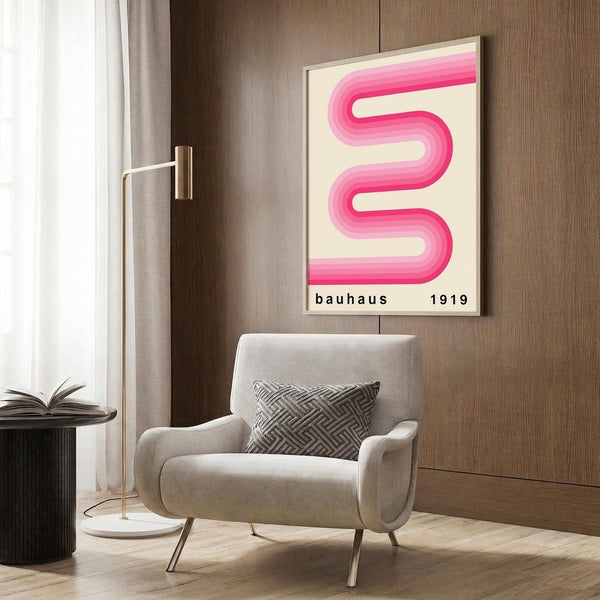 May include: Modern living room scene with beige armchair featuring geometric patterned pillow, gold floor lamp with white circular base, dark round side table with open book, and wood paneled wall displaying framed pink ribbon-shaped poster with gradient flowing curves and black text reading "bauhaus 1919" on cream background.