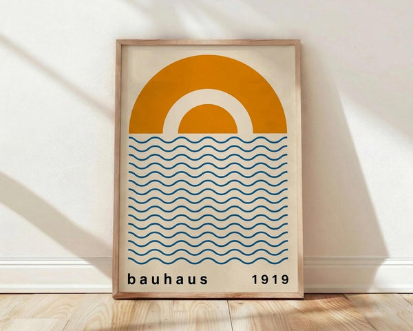 May include: Minimalist poster in light wood frame leaning against white wall on wooden floor. Features large orange semi-circle sun design above blue wavy lines representing water on cream background. Text reads "bauhaus 1919" in simple black lowercase letters. Natural lighting creates soft shadows on white textured wall.