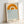 A minimalist poster in a light wood frame leaning against a white wall. The artwork features a geometric design with an orange double-arch sun above blue wavy horizontal lines representing water. Text at the bottom reads "bauhaus" and "1919" in simple black lettering. The poster has a cream-colored background and demonstrates clean Bauhaus design principles with bold, simple shapes and primary colors.