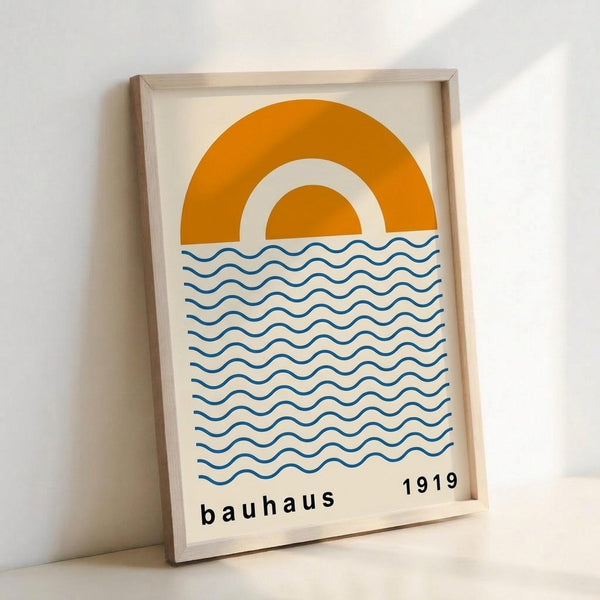 A minimalist poster in a light wood frame leaning against a white wall. The artwork features a geometric design with an orange double-arch sun above blue wavy horizontal lines representing water. Text at the bottom reads "bauhaus" and "1919" in simple black lettering. The poster has a cream-colored background and demonstrates clean Bauhaus design principles with bold, simple shapes and primary colors.
