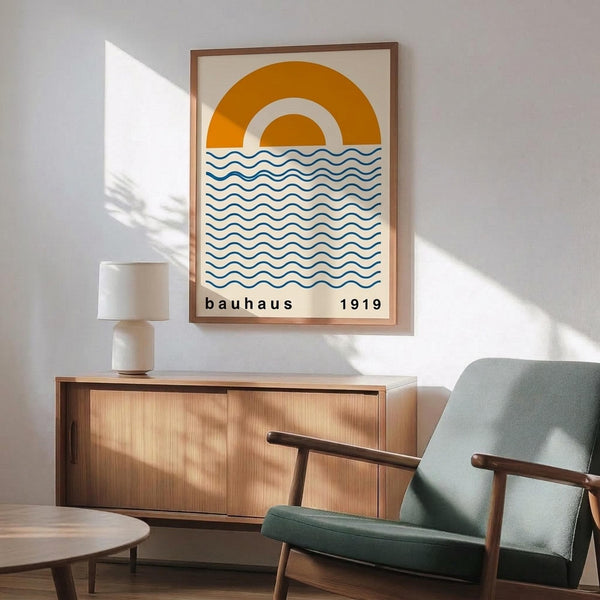 May include: Framed poster with orange and yellow concentric sun arcs above blue wavy lines on cream background, displayed above wooden sideboard with white ceramic lamp. Mid-century modern living room setting features sage green upholstered armchair with wooden frame, round wooden coffee table, white walls with dramatic sunlight shadows from window.