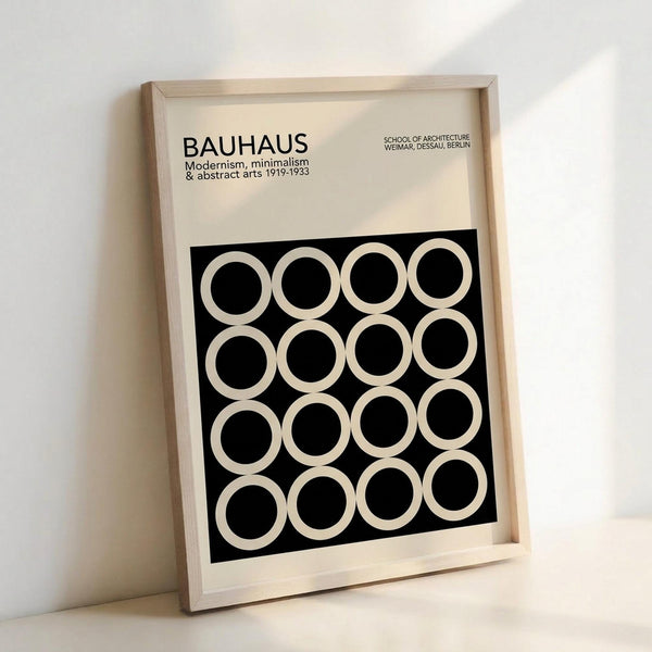 May include: Framed Bauhaus poster with cream background and black square containing 16 white circles arranged in 4x4 grid. Text reads "BAUHAUS Modernism, minimalism & abstract arts 1919-1933" and "SCHOOL OF ARCHITECTURE WEIMAR, DESSAU, BERLIN". Light wood frame leaning against white wall on white surface, creating soft shadows.