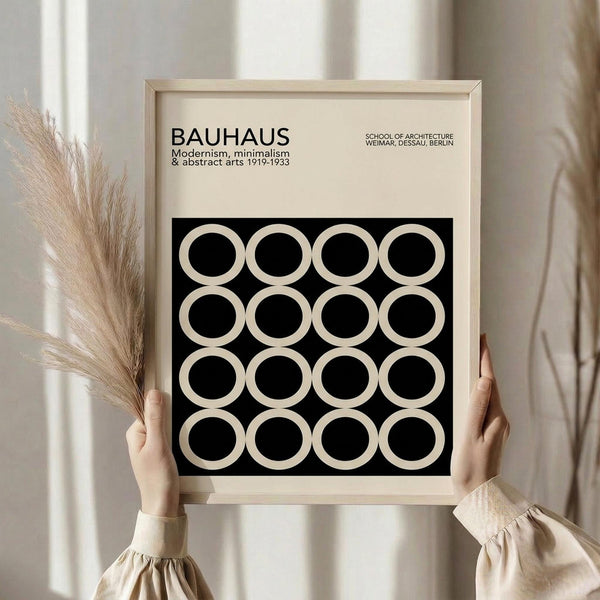 May include: Person in beige ruffled sleeve top holding framed Bauhaus poster against white curtained window. Poster features black background with 16 cream circles arranged in 4x4 grid, text about modernism and abstract arts 1919-1933. Light wooden frame, dried pampas grass visible on left side.