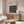 May include: Black and white Bauhaus poster with geometric circle pattern displayed above wooden sideboard in minimalist living room. White textured chair and glass coffee table visible. Poster features modernist design with repeating circular motifs on black background, framed in light wood. Natural lighting creates soft shadows across cream walls and warm wood furniture.