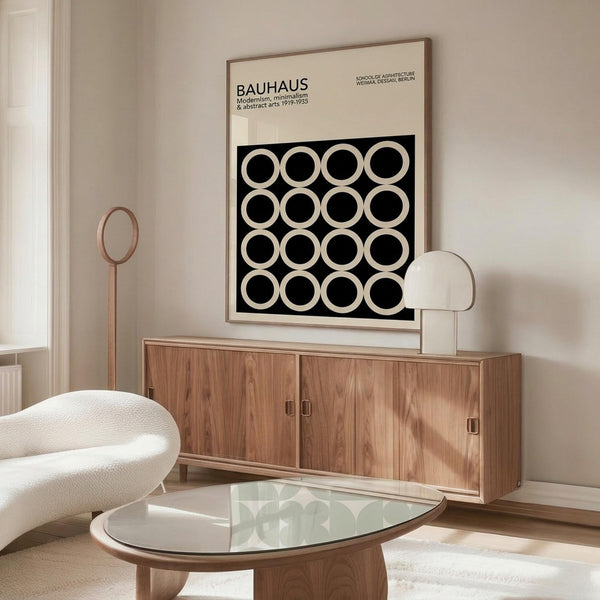 May include: Black and white Bauhaus poster with geometric circle pattern displayed above wooden sideboard in minimalist living room. White textured chair and glass coffee table visible. Poster features modernist design with repeating circular motifs on black background, framed in light wood. Natural lighting creates soft shadows across cream walls and warm wood furniture.