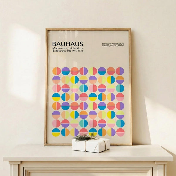 May include: Framed Bauhaus-inspired poster with colorful geometric circles in blues, yellows, oranges, and purples arranged in a grid pattern, displayed on light wood floating shelf against cream wall with small wrapped gift box beside it.
