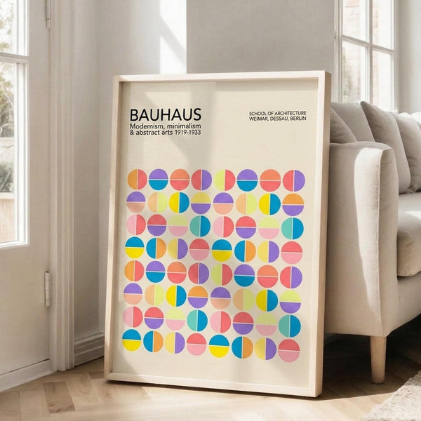 May include: A framed geometric poster with colorful circles split into halves in various bright colors including yellow, pink, blue, purple, orange, and green arranged in a grid pattern. The poster features "BAUHAUS" text at top and sits in a light wood frame leaning against a white wall next to a beige upholstered chair in a bright, naturally-lit room with white walls and wood floors.