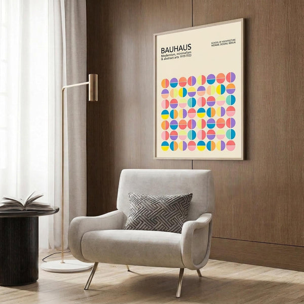 May include: Framed Bauhaus poster with colorful geometric circles in pink, blue, yellow, and orange displayed on wood paneled wall above beige upholstered armchair with patterned throw pillow. Modern brass floor lamp and dark round side table with open book complete the minimalist room setting with light wood floors.