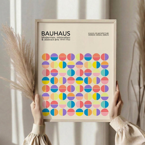 May include: Person holding a framed Bauhaus-inspired poster featuring colorful geometric circles in a grid pattern. The artwork displays vibrant blues, oranges, purples, pinks, and yellows on a cream background with "BAUHAUS" text at the top. Natural wood frame, soft neutral room setting with pampas grass visible, hands wearing cream-colored sleeves with gathered cuffs.
