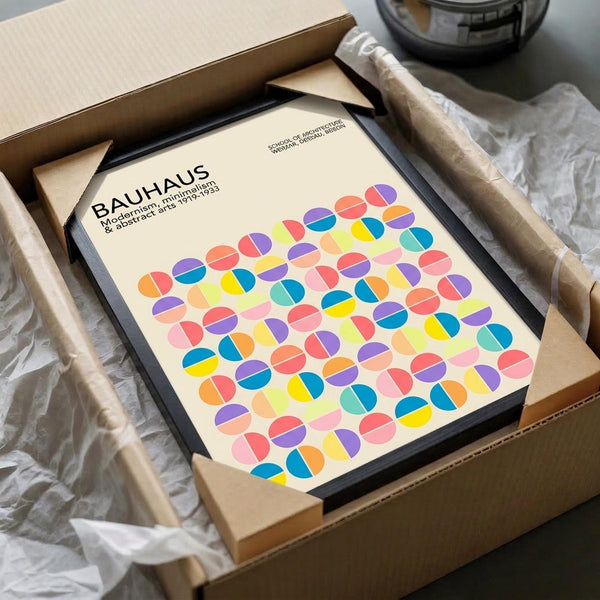 May include: Framed Bauhaus poster with colorful geometric circles in a grid pattern, featuring vibrant blues, yellows, pinks, and purples on cream background. Black frame nestled in cardboard packaging with white tissue paper, sitting on white fabric surface next to partial view of glass bowl.