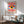 May include: Framed Bauhaus-style poster featuring a stylized eye with yellow iris, dark blue pupil, and white highlight, surrounded by red and white radiating sunburst pattern on cream background with blue accents. Displayed above modern white sideboard with ceramic lamp, next to mid-century green upholstered armchair with wooden frame in bright, naturally-lit room with white walls.