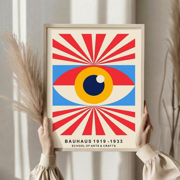 May include: Person holding a framed Bauhaus-style poster featuring a stylized eye with blue iris, yellow surrounding area, and red radiating sunburst pattern against cream background. Light wood frame, neutral room setting with dried pampas grass visible to the left.