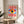 May include: Framed Bauhaus-inspired poster featuring a stylized eye with yellow iris, blue pupil, and white highlight, surrounded by radiating red and white sunburst rays on cream background. The eye sits within a blue rectangular frame. Print displayed in thin wooden frame on round wooden side table next to beige upholstered chair, with plant leaves visible in soft natural lighting.
