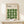 May include: Framed poster with green geometric pattern made of curved leaf-like shapes arranged in a grid, displayed on light wood shelf next to small wrapped gift with twine and evergreen sprig. Poster has cream background with black text reading "BAUHAUS" and details about modernism and abstract arts. Natural wood frame against neutral wall.