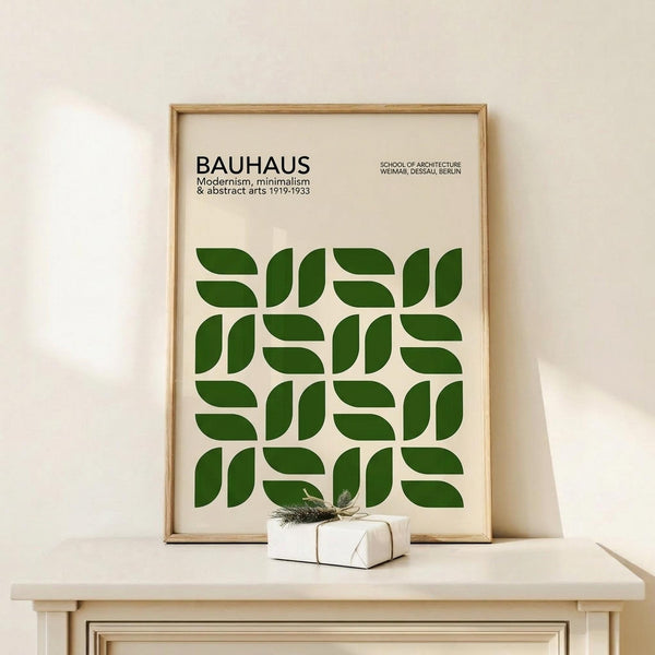 May include: Framed poster with green geometric pattern made of curved leaf-like shapes arranged in a grid, displayed on light wood shelf next to small wrapped gift with twine and evergreen sprig. Poster has cream background with black text reading "BAUHAUS" and details about modernism and abstract arts. Natural wood frame against neutral wall.