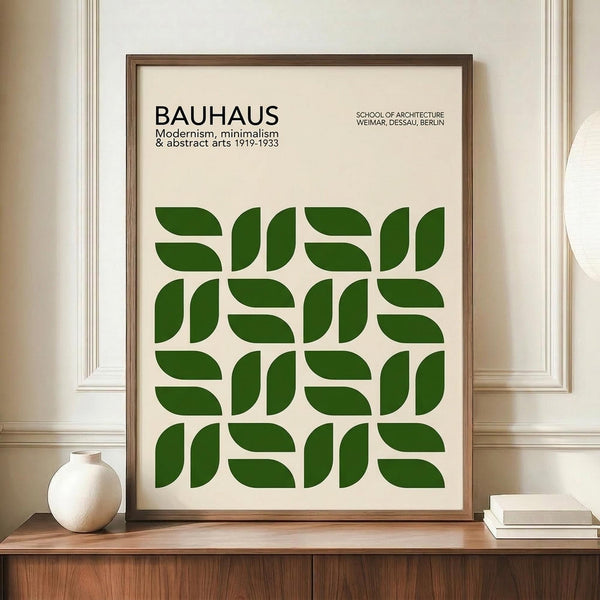 May include: Framed poster with cream background featuring green geometric leaf-like shapes arranged in a 4x4 grid pattern, with "BAUHAUS" text at top and "Modernism, minimalism & abstract arts 1919-1933" below, plus "School of Architecture Weimar, Dessau, Berlin" in upper right. Natural wood frame sits on wooden sideboard next to white ceramic vase and stacked books against white paneled wall.