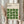 May include: Person in beige blouse holding framed Bauhaus poster with geometric green leaf pattern on cream background. Light wood frame, dried pampas grass visible to left, soft natural lighting by window with white curtains.