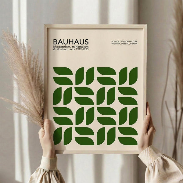 May include: Person in beige blouse holding framed Bauhaus poster with geometric green leaf pattern on cream background. Light wood frame, dried pampas grass visible to left, soft natural lighting by window with white curtains.