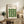 May include: Framed Bauhaus poster with green geometric leaf pattern displayed on rustic wooden sideboard next to fern plant, books, and ceramic vase in neutral-toned living room setting with beige upholstered chair