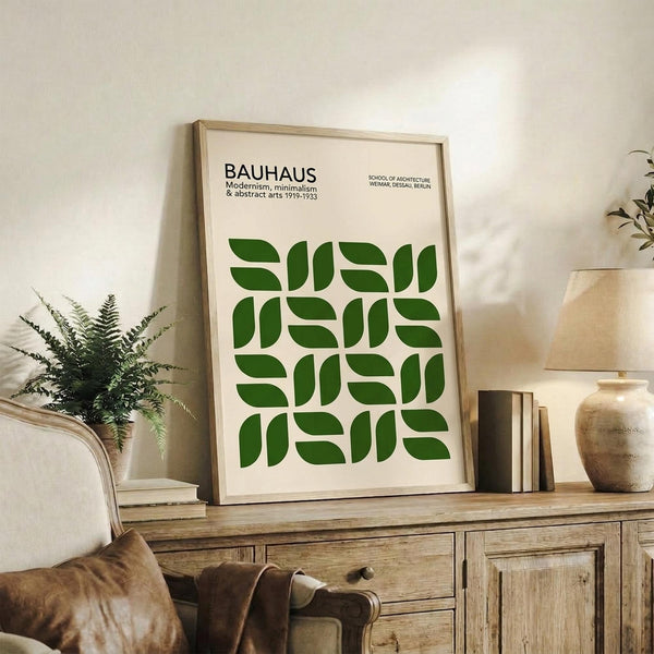 May include: Framed Bauhaus poster with green geometric leaf pattern displayed on rustic wooden sideboard next to fern plant, books, and ceramic vase in neutral-toned living room setting with beige upholstered chair