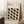 May include: Framed Bauhaus poster with geometric leaf pattern in black, olive green, and sage colors leaning against white wall. Modern minimalist design with text about 1919-1933 modernism movement. Natural wood frame, positioned next to wooden block with white coffee cup, cream textured rug, and beige throw blanket visible in corner.