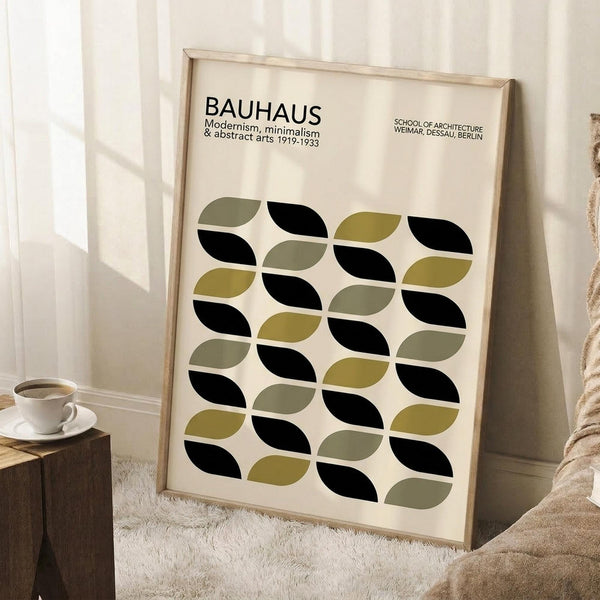 May include: Framed Bauhaus poster with geometric leaf pattern in black, olive green, and sage colors leaning against white wall. Modern minimalist design with text about 1919-1933 modernism movement. Natural wood frame, positioned next to wooden block with white coffee cup, cream textured rug, and beige throw blanket visible in corner.
