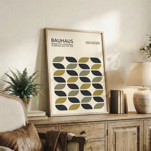 May include: Framed Bauhaus poster with geometric leaf pattern in olive green and dark teal on cream background, displayed on rustic wooden sideboard alongside fern plant, ceramic vases, and leather armchair in neutral-toned living room setting