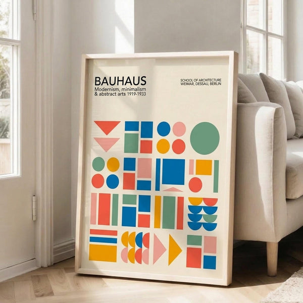 May include: Framed Bauhaus poster with colorful geometric shapes including triangles, circles, squares and rectangles in red, blue, green, orange and pink on cream background. Light wood frame leans against white wall next to beige upholstered chair in bright room with tall windows and hardwood floors.