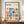 May include: Framed Bauhaus poster with colorful geometric shapes - triangles, circles, squares and rectangles in blue, orange, red, pink and green arranged in a grid pattern. Text reads "BAUHAUS Modernism, minimalism & abstract arts 1919-1933" and "School of Architecture Weimar, Dessau, Berlin". Brown wooden frame displayed on wooden sideboard next to white ceramic vase and books against cream wall with white trim molding.