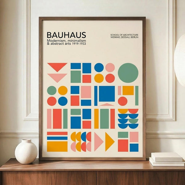 May include: Framed Bauhaus poster with colorful geometric shapes - triangles, circles, squares and rectangles in blue, orange, red, pink and green arranged in a grid pattern. Text reads "BAUHAUS Modernism, minimalism & abstract arts 1919-1933" and "School of Architecture Weimar, Dessau, Berlin". Brown wooden frame displayed on wooden sideboard next to white ceramic vase and books against cream wall with white trim molding.