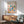 May include: Framed Bauhaus poster with colorful geometric shapes - triangles, circles, rectangles in blue, red, yellow, green, and pink - displayed above a mid-century modern wooden credenza with white ceramic lamp. Sage green upholstered chair with wooden arms sits beside round coffee table. Bright natural light casts shadows on white wall.