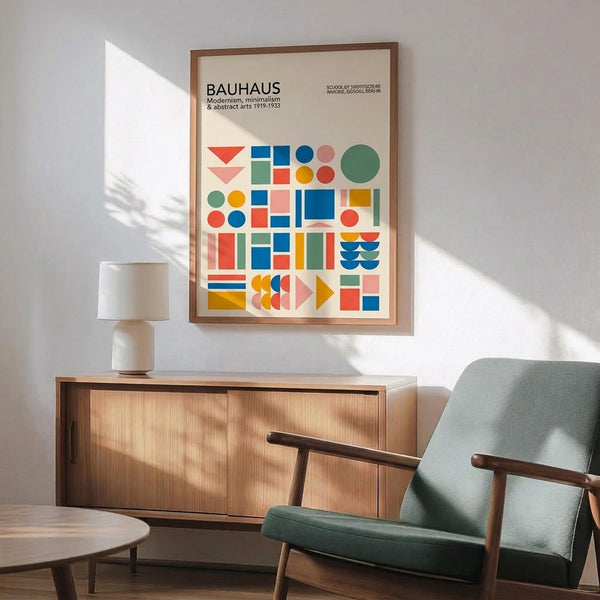 May include: Framed Bauhaus poster with colorful geometric shapes - triangles, circles, rectangles in blue, red, yellow, green, and pink - displayed above a mid-century modern wooden credenza with white ceramic lamp. Sage green upholstered chair with wooden arms sits beside round coffee table. Bright natural light casts shadows on white wall.