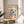 May include: Framed Bauhaus poster with colorful geometric shapes - circles, squares, triangles, and rectangles in red, blue, yellow, green, and pink - displayed on round wooden side table next to beige upholstered chair. Natural light streams across white wall creating soft shadows. Plant leaves visible in corner of minimalist, modern interior setting.