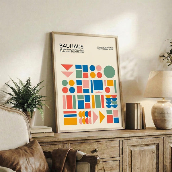 May include: Framed Bauhaus poster with colorful geometric shapes in red, blue, green, orange, and pink displayed on rustic wooden sideboard alongside fern plant, ceramic vase, and books in neutral-toned living room setting with beige upholstered furniture