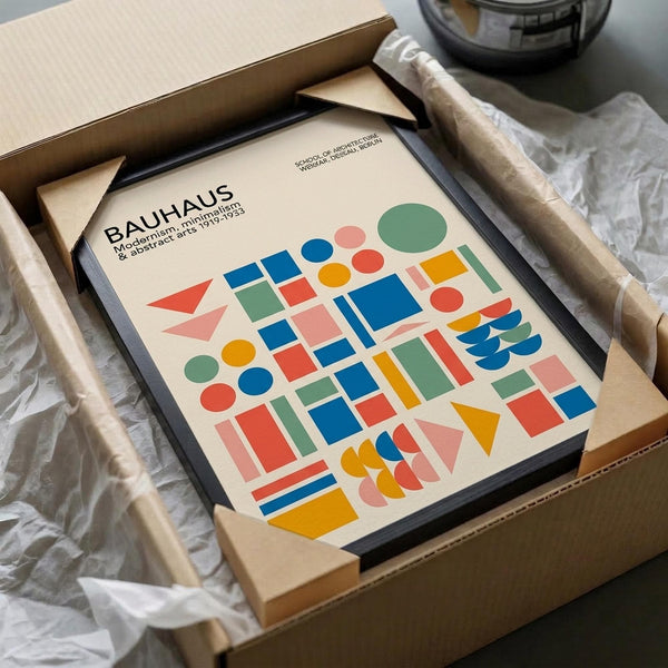 May include: Framed Bauhaus poster with colorful geometric shapes including circles, squares, triangles and rectangles in blue, red, green, yellow, pink and orange on cream background. Black frame sits in cardboard shipping box with white tissue paper padding on wooden surface.