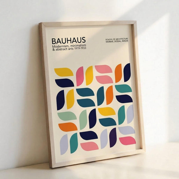 Framed poster with cream background featuring colorful geometric half-circle and oval shapes in yellow, pink, coral, navy, teal, orange, and lavender. Black text at top reads "BAUHAUS Modernism, minimalism & abstract arts 1919-1933" and "SCHOOL OF ARCHITECTURE WEIMAR, DESSAU, BERLIN." Light wood frame sits against white wall casting soft shadows.