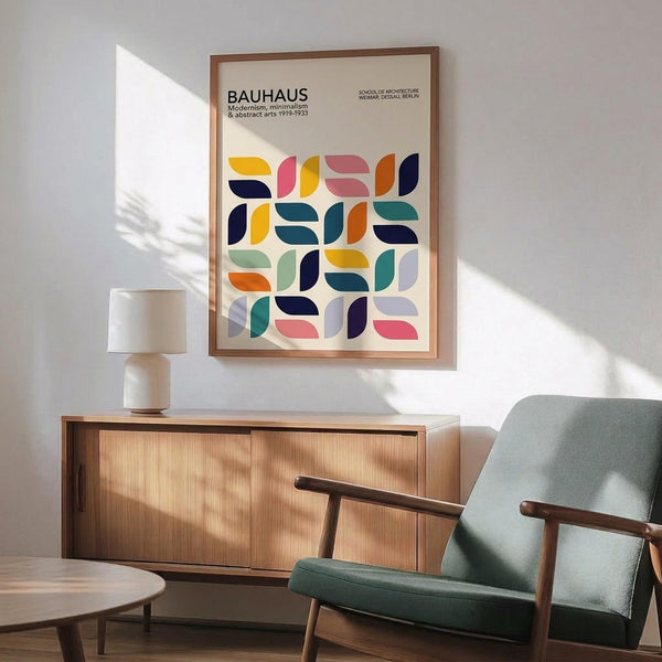May include: Modernist poster with colorful geometric half-circle and leaf shapes in yellow, coral, navy, teal, and orange arranged in a grid pattern, displayed in wooden frame above mid-century credenza with table lamp, next to sage green upholstered armchair with wooden arms in sunlit room with white walls