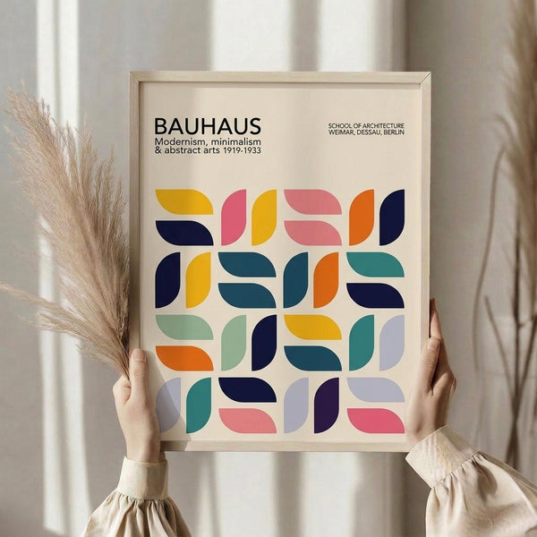 May include: Person holding framed Bauhaus poster featuring colorful geometric semicircle pattern in navy, yellow, pink, orange, teal and mint green. Light wood frame against cream background with dried pampas grass stems visible. Minimalist modernist design with clean typography referencing the architecture school's 1919-1933 period.
