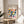 May include: Framed Bauhaus poster with colorful geometric half-circle pattern in pink, yellow, orange, teal, navy and green on cream background, displayed on light wood round side table next to beige upholstered chair with plant leaves visible, natural sunlight creating shadows on white wall