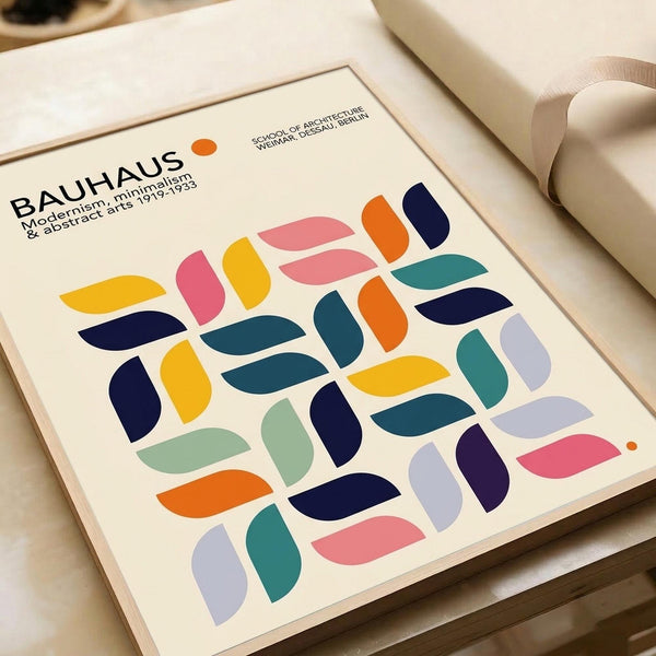 May include: Framed Bauhaus poster print with colorful geometric leaf-like shapes in pink, yellow, orange, navy, teal, and purple arranged in a grid pattern. The cream-colored print features "BAUHAUS" text and "Modernism, minimalism & abstract arts 1919-1933" with "School of Architecture, Weimar, Dessau, Berlin" subtitle. Frame sits on white bedding next to a beige pillow, showing the minimalist mid-century modern design aesthetic.