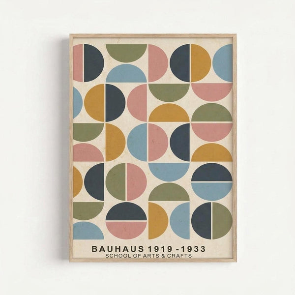 Framed geometric poster featuring colorful semicircles and quarter-circles arranged in a grid pattern. The artwork displays muted tones of coral pink, sage green, mustard yellow, dusty blue, and charcoal gray on a cream background. Text at bottom reads "BAUHAUS 1919-1933 SCHOOL OF ARTS & CRAFTS" in black letters. The print is mounted in a simple light wood frame and hung on a white wall.